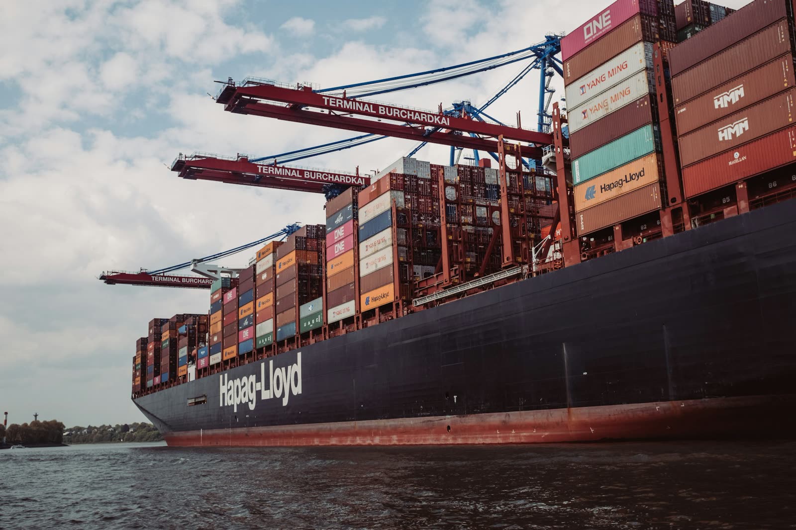 The bow of a container vessel with Hapag-Lloyd on its hull, alongside at a terminal.