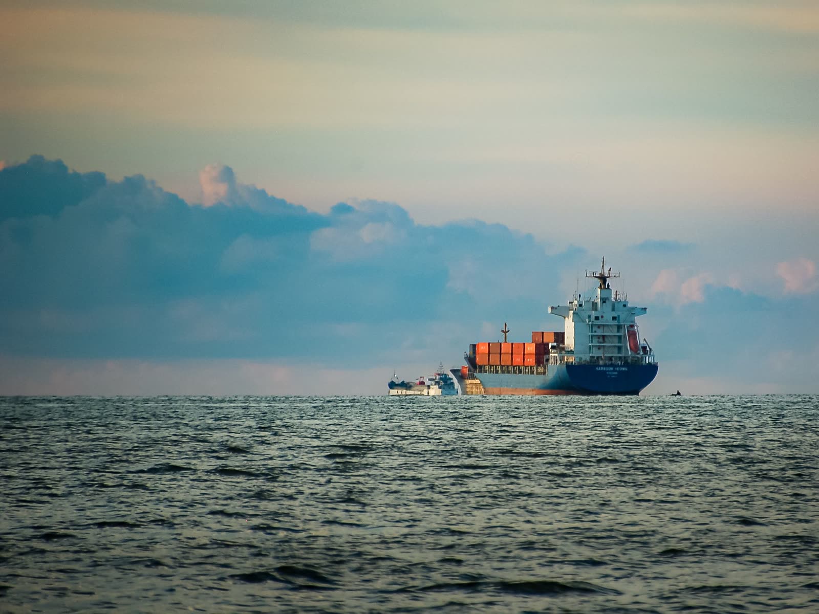 A small container vessel far off on open water at dusk, under a bank of cloud.
